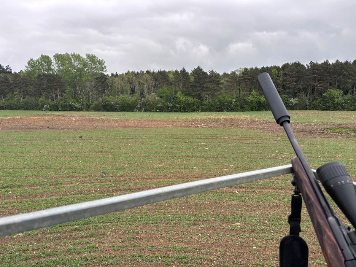 A suppressed rifle resting on a field gate, overlooking farmland bordered by woodland under an overcast Yorkshire sky